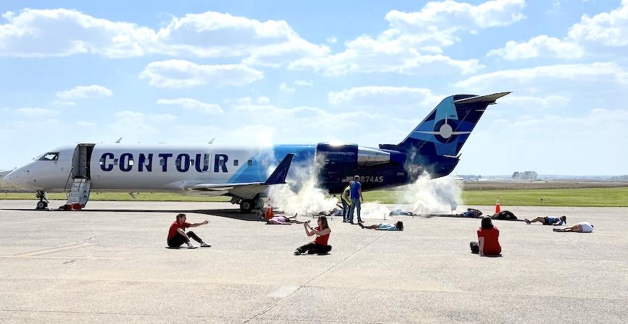 A CONTOUR-branded airplane is parked on a runway with smoke coming from its rear. Several people, some in red shirts, are lying on the ground around the plane in various positions. The scene appears to depict a simulated emergency or training exercise. The sky is mostly clear with scattered clouds, and grassy fields are visible in the background.