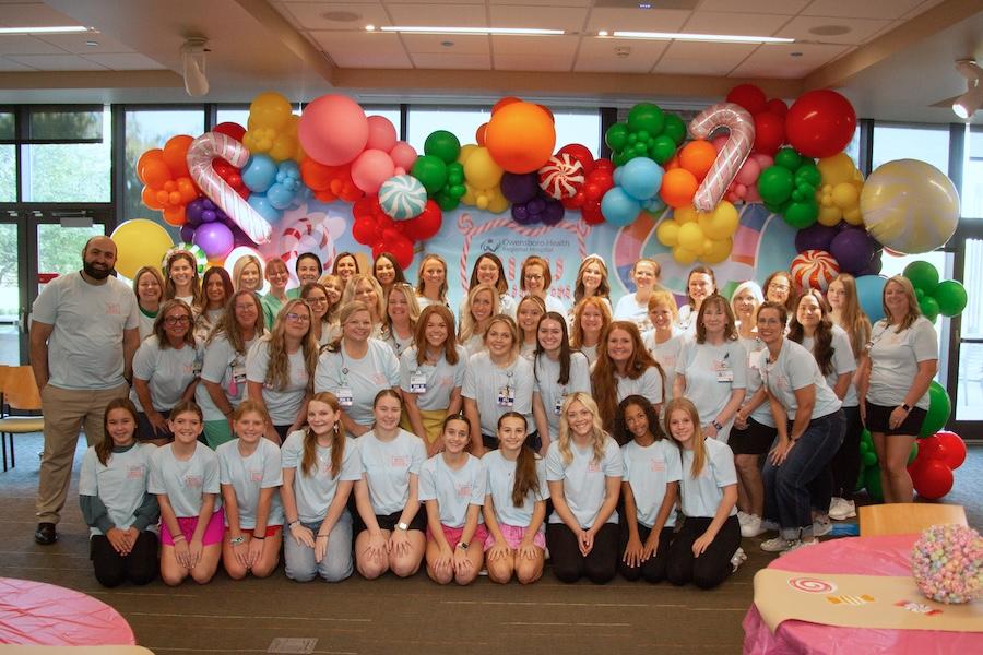 A large group of people in light blue shirts pose for a group photo in a festively decorated room with colorful balloon arches, candy-themed inflatables, and tables with pink tablecloths and lollipop centerpieces.